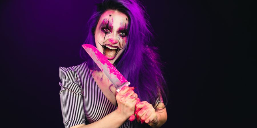 Halloween party ideas for adults featuring a woman in creepy clown makeup holding a bloody knife prop for a scary costume theme.