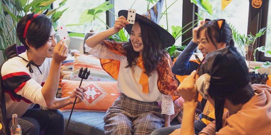 Halloween party games with friends in costumes laughing and playing cards together at a festive celebration.