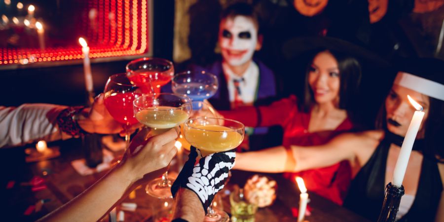 Halloween party ideas for adults with friends in costumes raising colorful cocktails in a festive toast by candlelight.