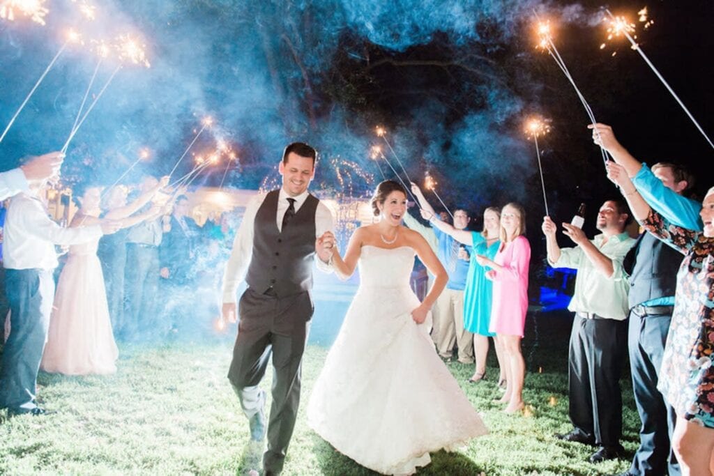 A gleeful bride and groom run through a group of friends holding tall sparklers