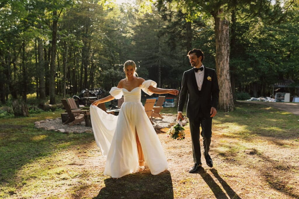 A bride laughs while she walks alongside her groom in a wooded area while holding up the train of her dress as he smiles at her while holding her bouquet