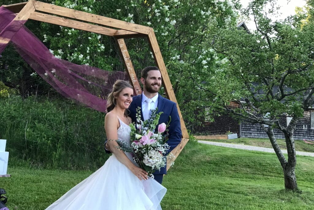 A bride and groom stand by a wood altar draped in purple tulle outside by a clapboard house