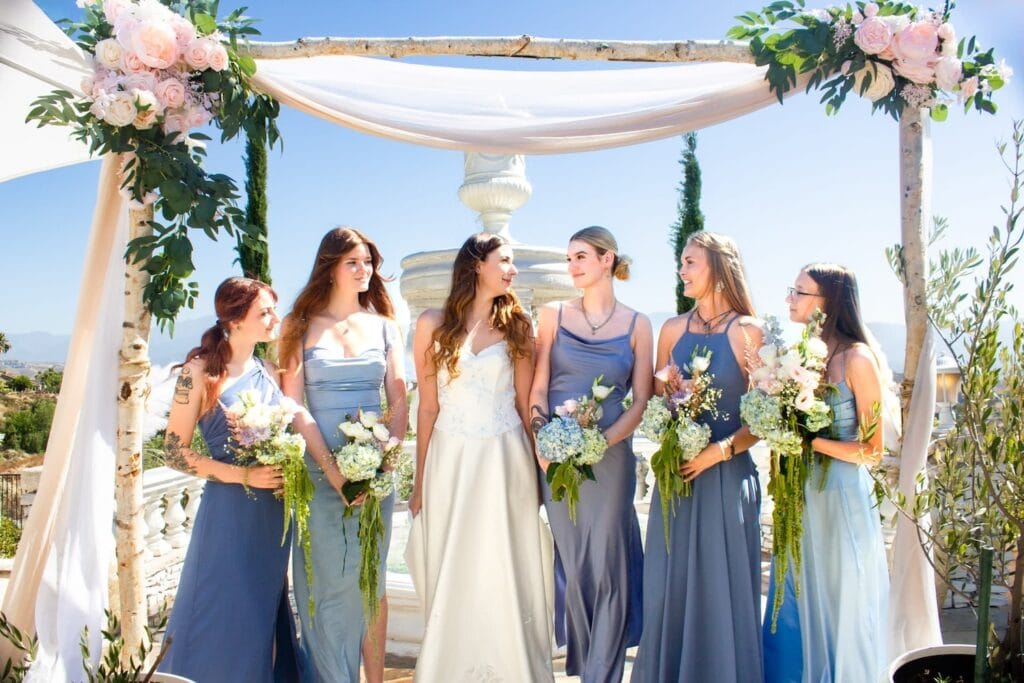 Bridesmaids in blue surround a bride outside under an archway in the sunshine.