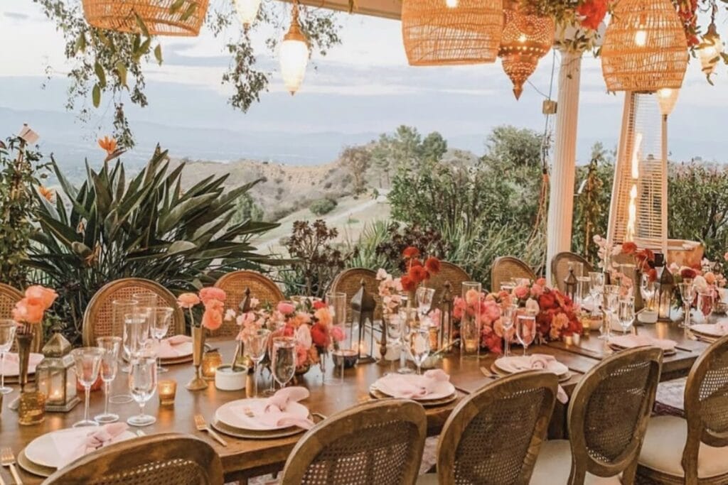 A long reception table is set with low-profile pink and peach centerpieces on a veranda overlooking hills