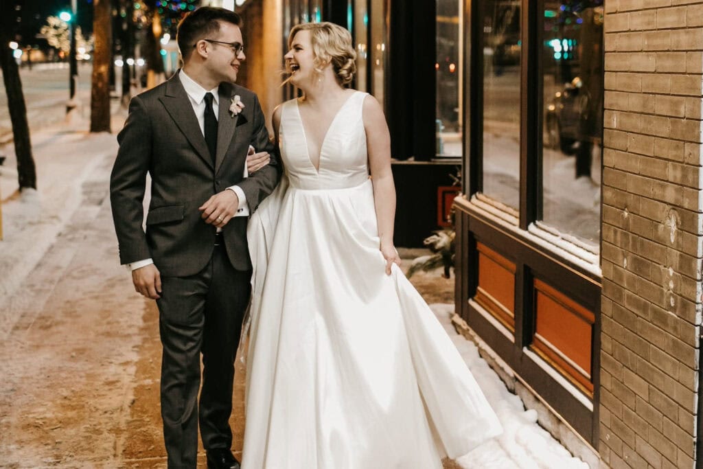 A bride and groom walk arm in arm down a snowy street laughing together