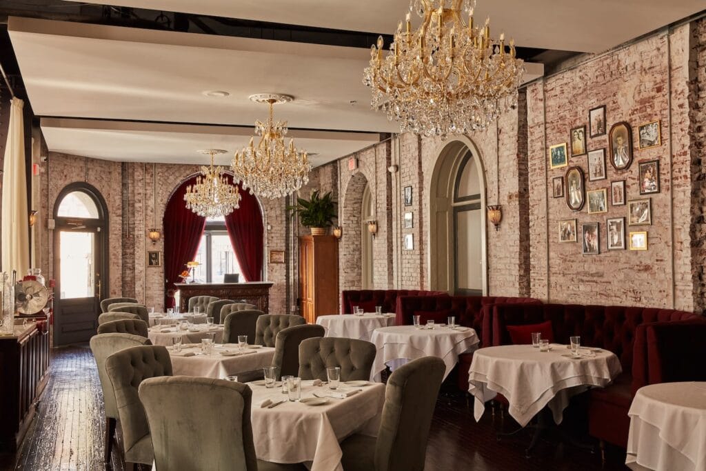 An historic restaurant space with high ceilings, exposed brick walls, elaborate chandeliers and red and green velvet seating