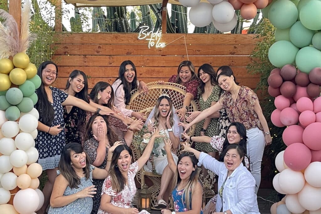 A bride to be is surrounded by a bunch of friends and family outside with balloons and flowers to mark the occasion