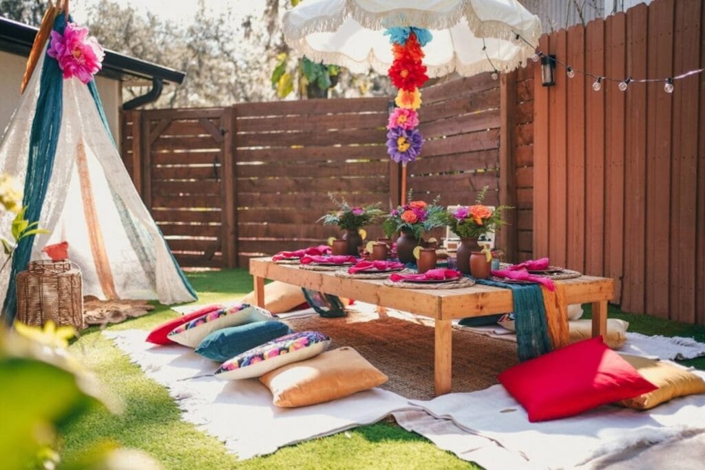 Bright flowers line a low-lying table surrounded by colorful pillows in a sunny backyard area next to a teepee
