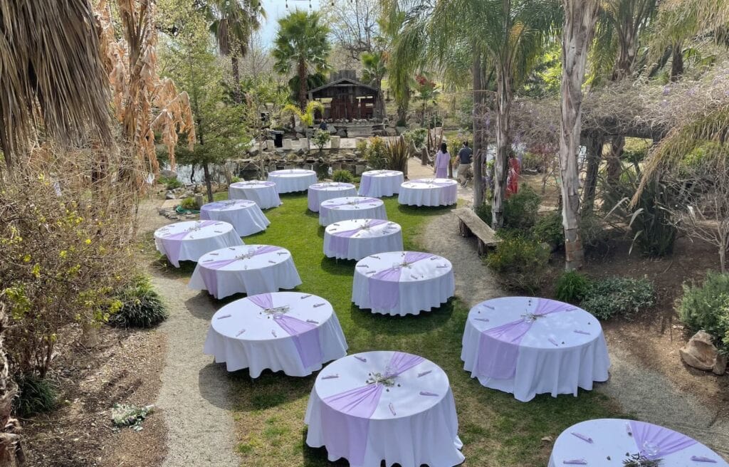Round banquet tables are set up outside in a clearing at a rustic venue surrounded by lush greenery