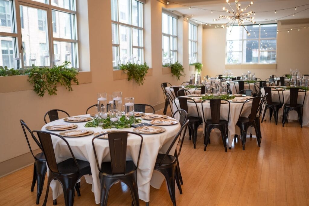 A bright, white event space lit by a bank of large windows is set up with round tables and metal chairs for a small reception