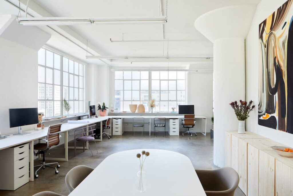 An all-white workspace with large windows looking out onto a city and long white desks with different style desk chairs and beautiful art on the walls