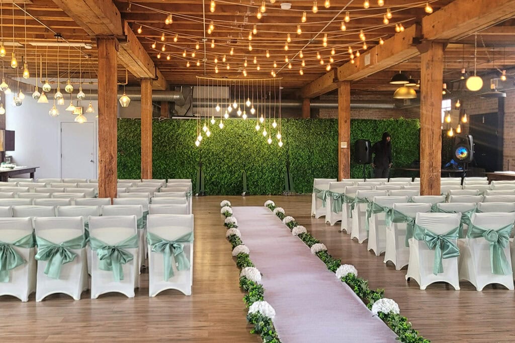 Indoor Dallas wedding ceremony setup with white chairs, green ribbons, leafy backdrop, and string lights