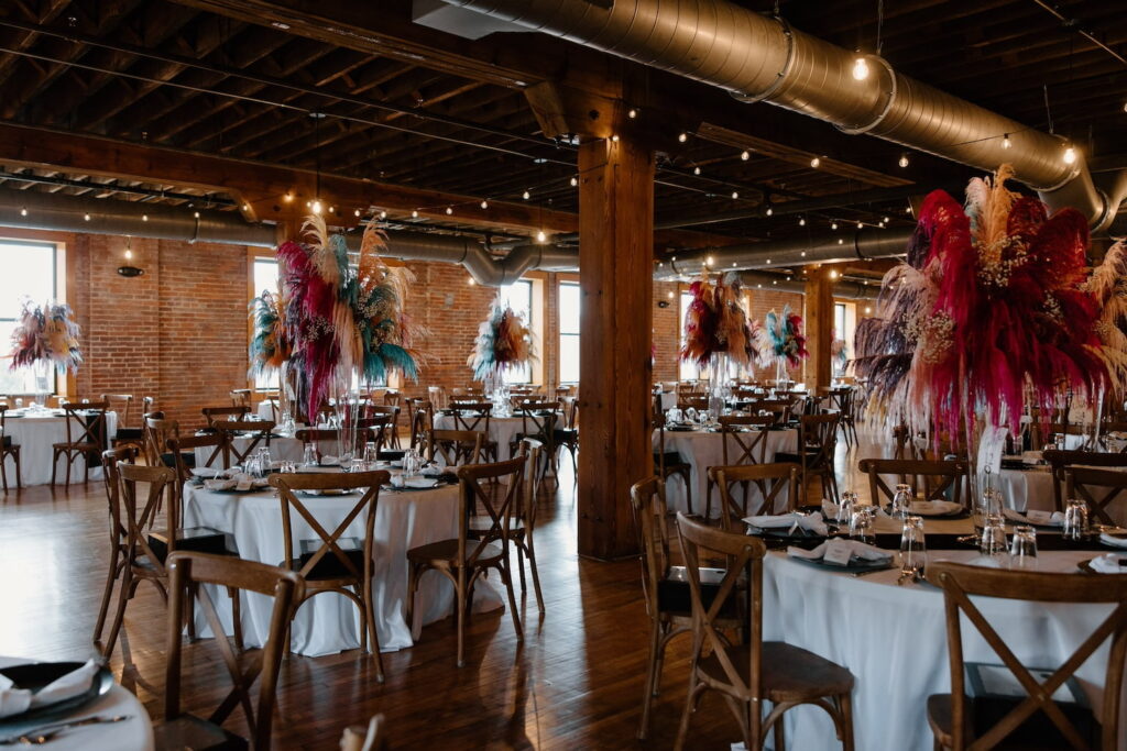 Dramatic centerpieces of boldly-colored feathers decorate round tables in a rustic reception venue with brick walls and exposed wooden beams