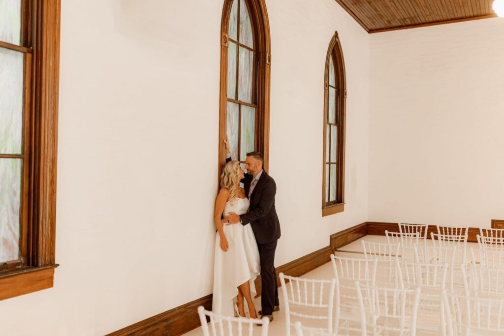 A bride and groom steal a quiet moment by a large wooden window in an all-white ceremony venue with white chairs
