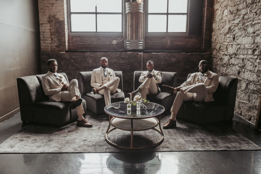 Groomsmen sit facing the camera in an industrial venue with large windows and stone walls