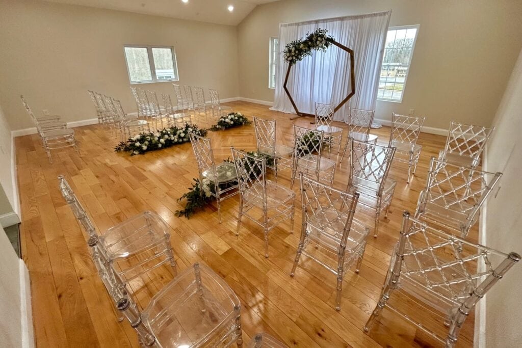 Clear chairs are set up in a small room arranged for a wedding ceremony with flowers lining the aisle and a wooden altar draped in greenery