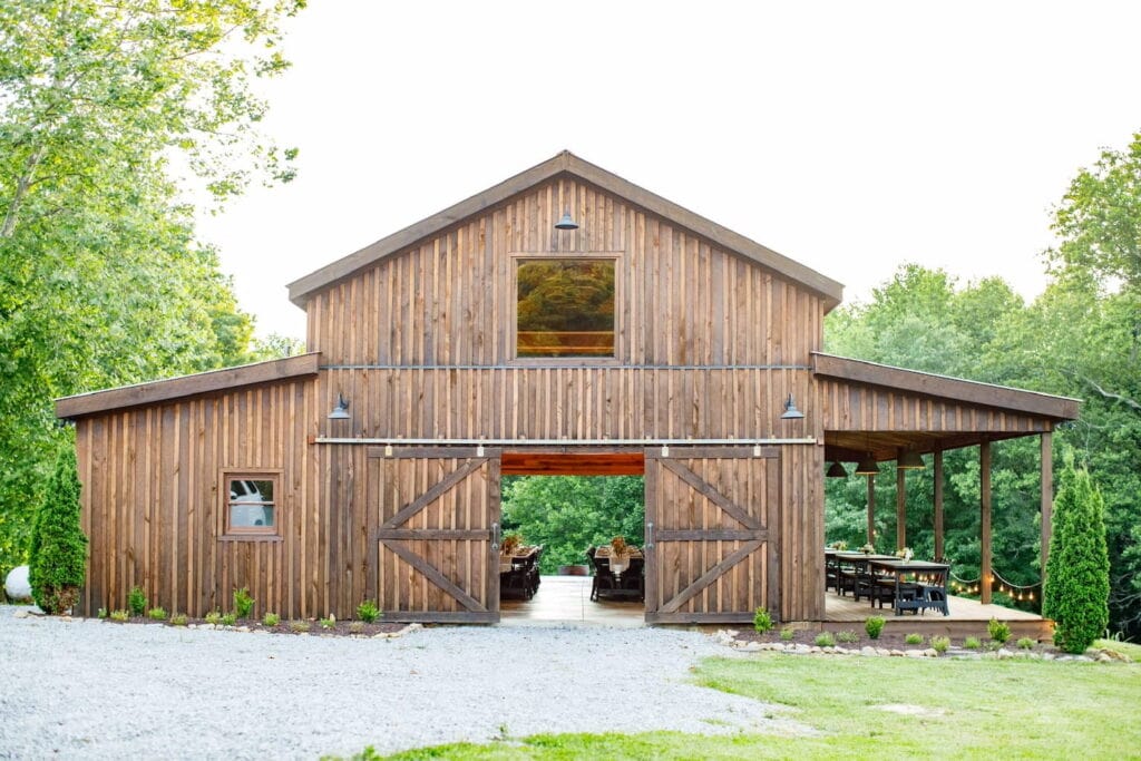 A large wooden barn sits in the woods set up for a wedding reception inside the barn doors and outside on a covered deck
