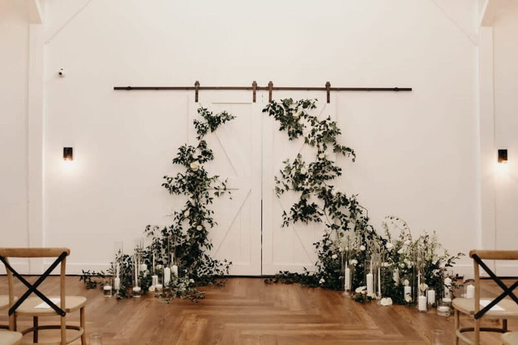 White barn doors are the backdrop for a simple wedding altar decorated in greenery and surrounded by candles in an all-white venue