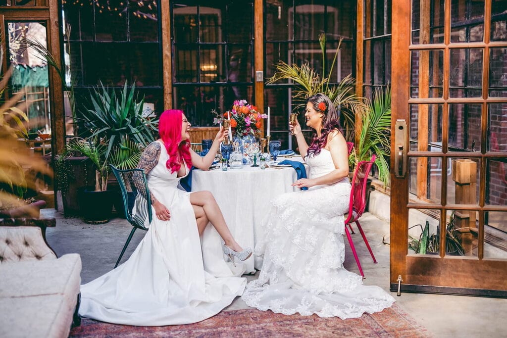 Two brides hold champagne glasses while smiling at each other at a table decorated with brightly-colored flowers and blue accents