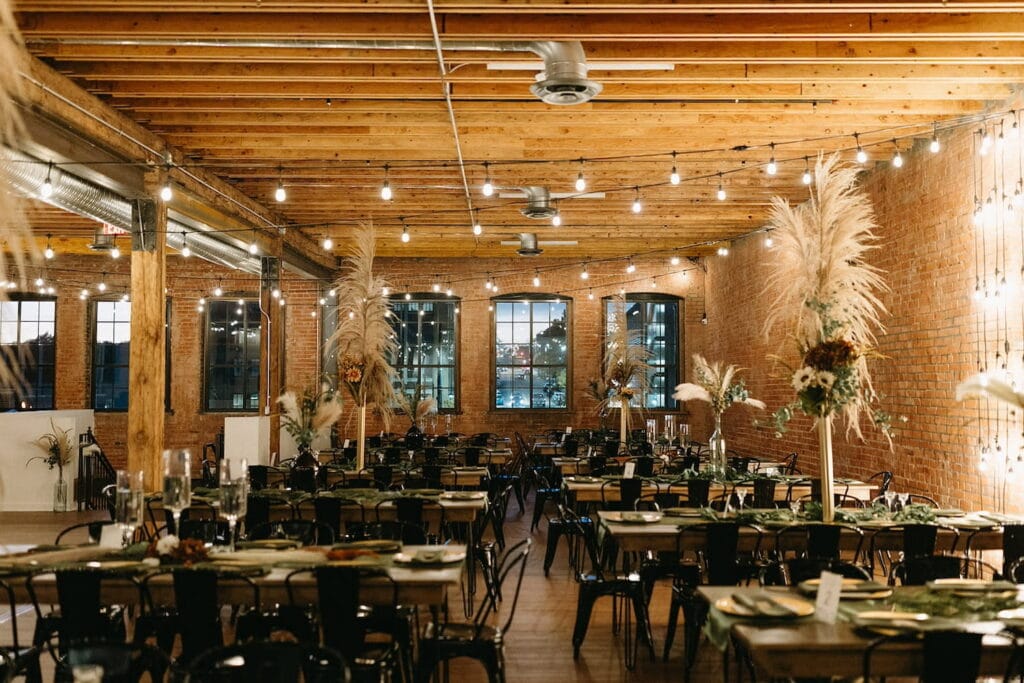 Exposed brick and wooden beams provide a warm backdrop for a beautifully lit reception space decorated with wooden tables, black chairs and tall centerpieces