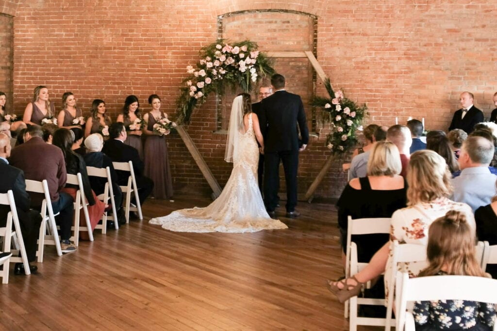 A bride and groom stand at an altar made of wood and draped in flowers surrounded by friends and family in a rustic event venue