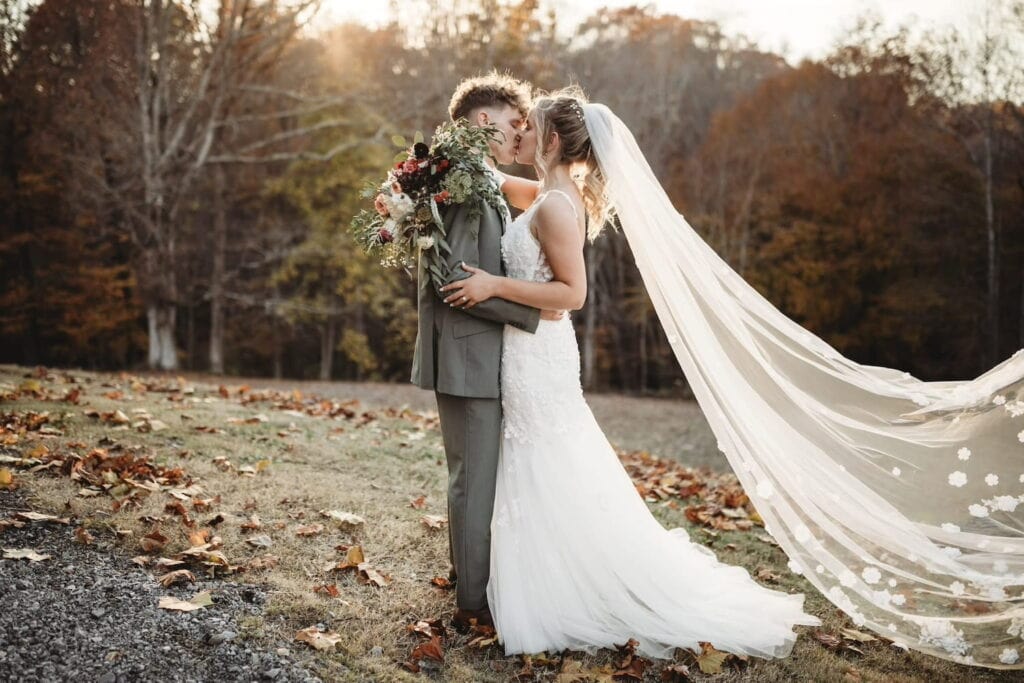 A bride and groom kiss in the woods with the sun setting behind them