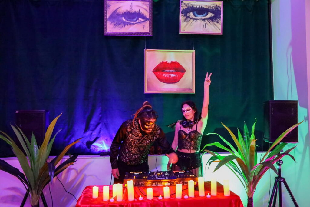 Two DJs in masks stand at a turntable at a party space beneath hanging art