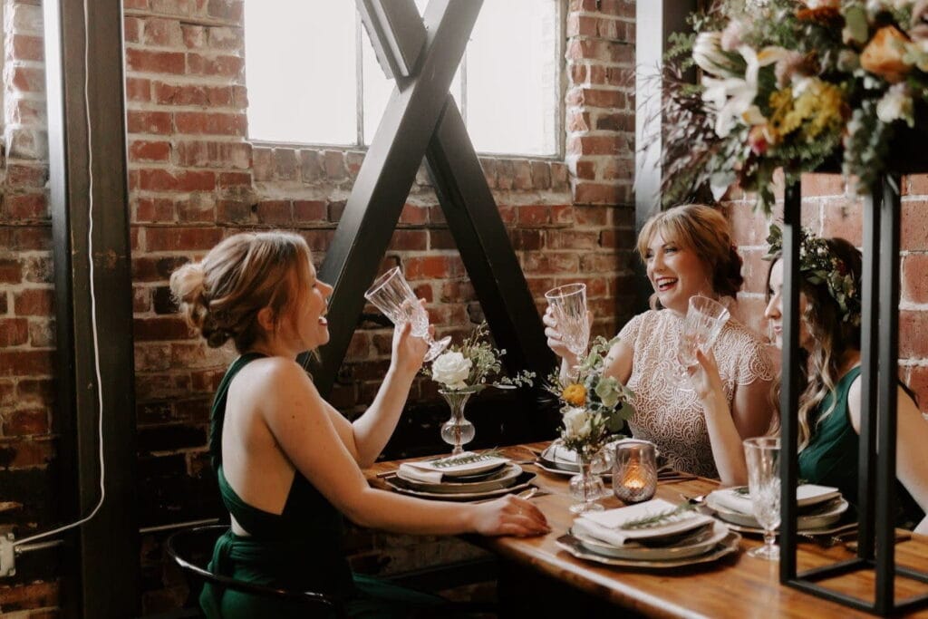 A bride toasts with two women at a table in an industrial space with brick walls