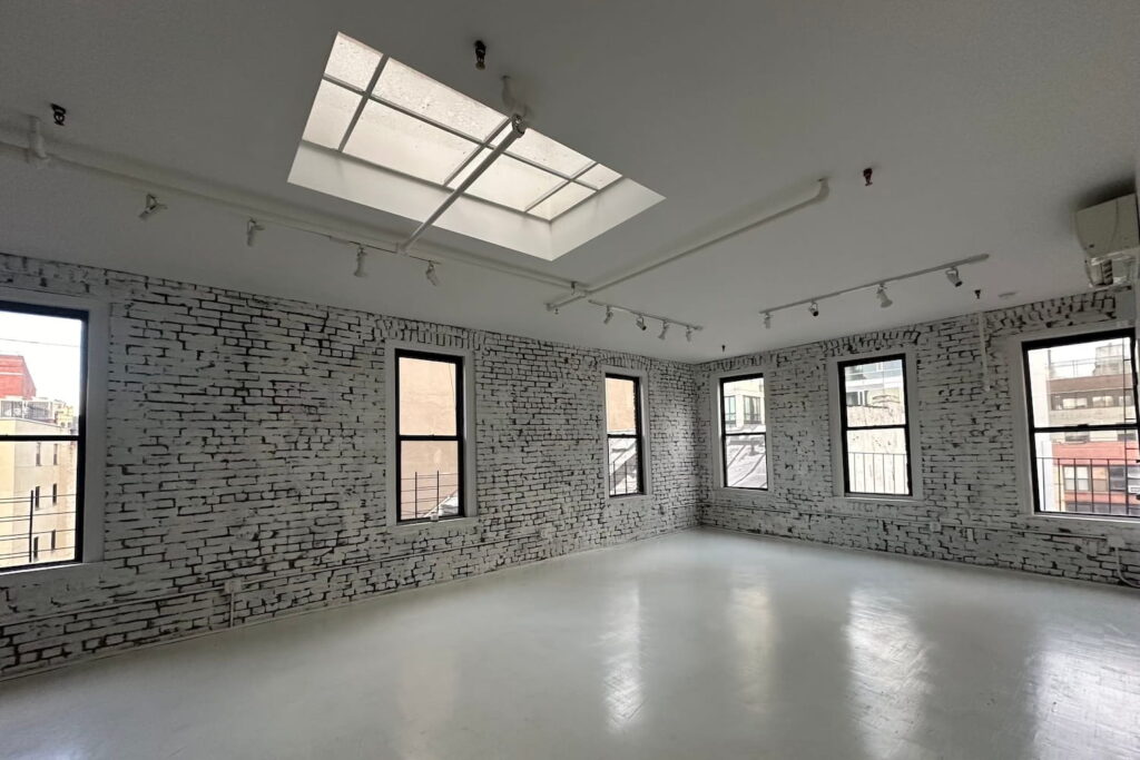 Large windows and a skylight illuminate a large open event space with painted brick walls and white cement floor