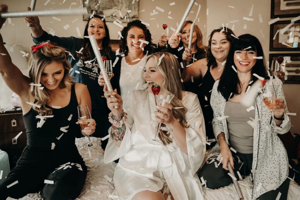 A bride is surrounded by her bridal squad as they hold champagne glasses and confetti falls around them