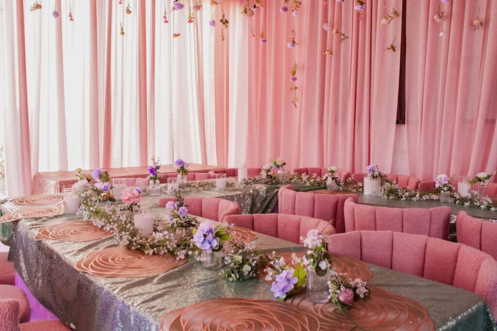 All pink party space with pink upholstered chairs around a U-shaped banquet table covered in gold cloth and purple flowers