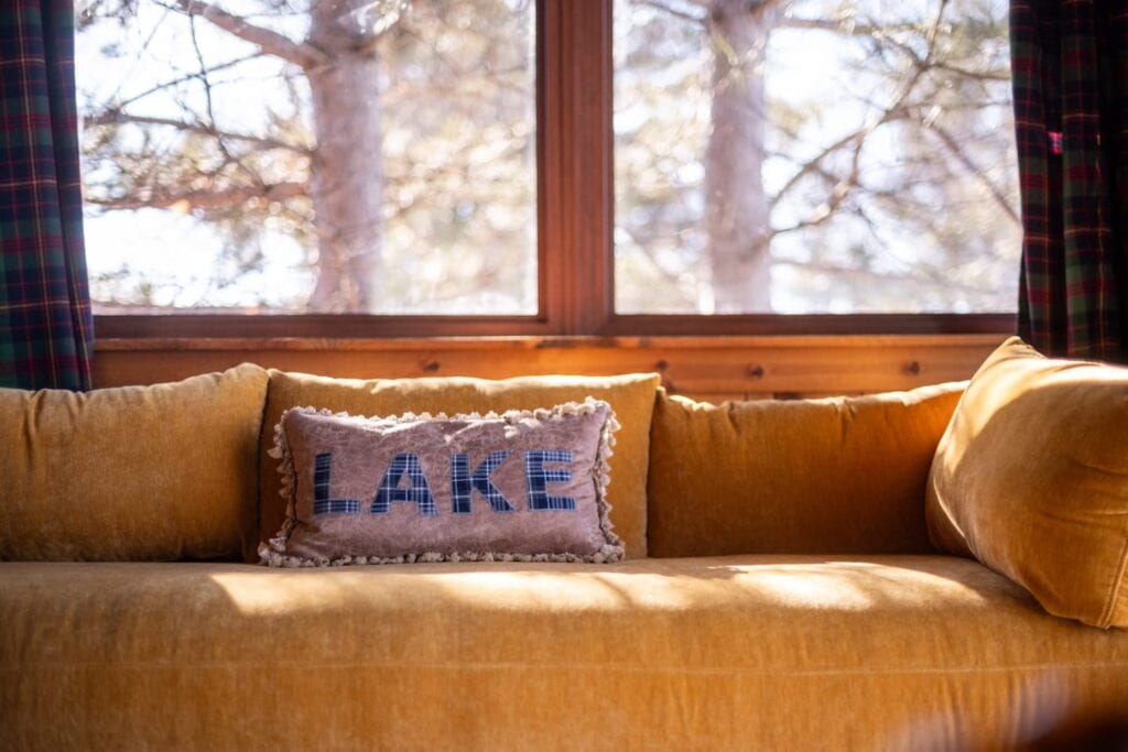 A pillow with the word "Lake" embroidered on it sits on a yellow couch by a large window looking out into the woods