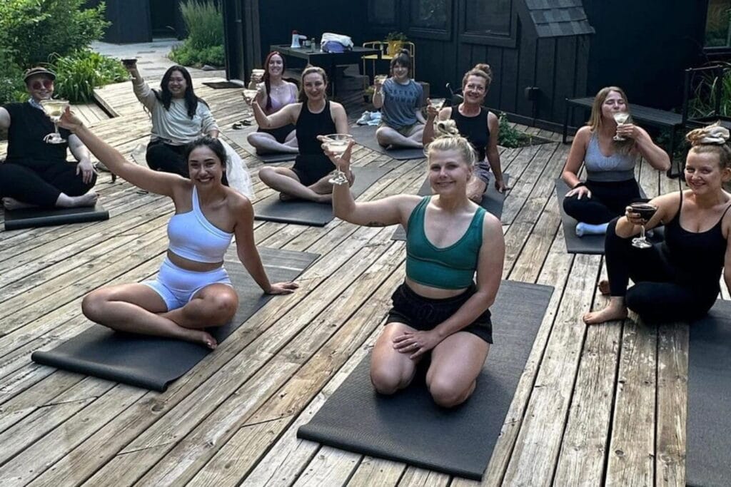 A group of women holding martini glasses while on yoga mats outside on a deck with a woman in the front in all white and a veil