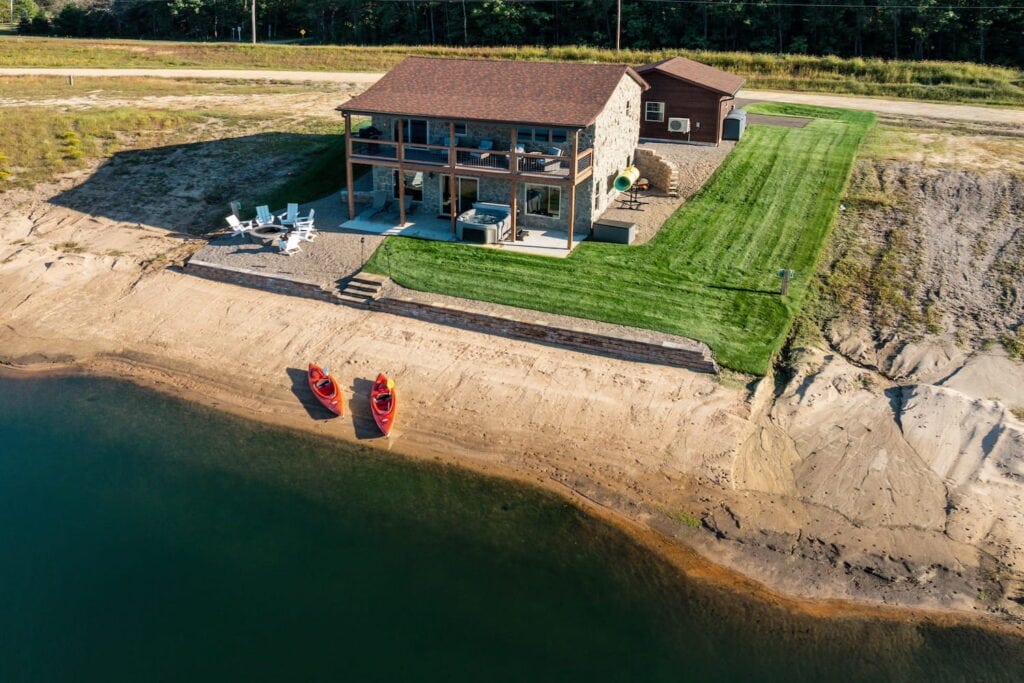 Two kayaks sit on a lake shoreline outside of a large waterfront home