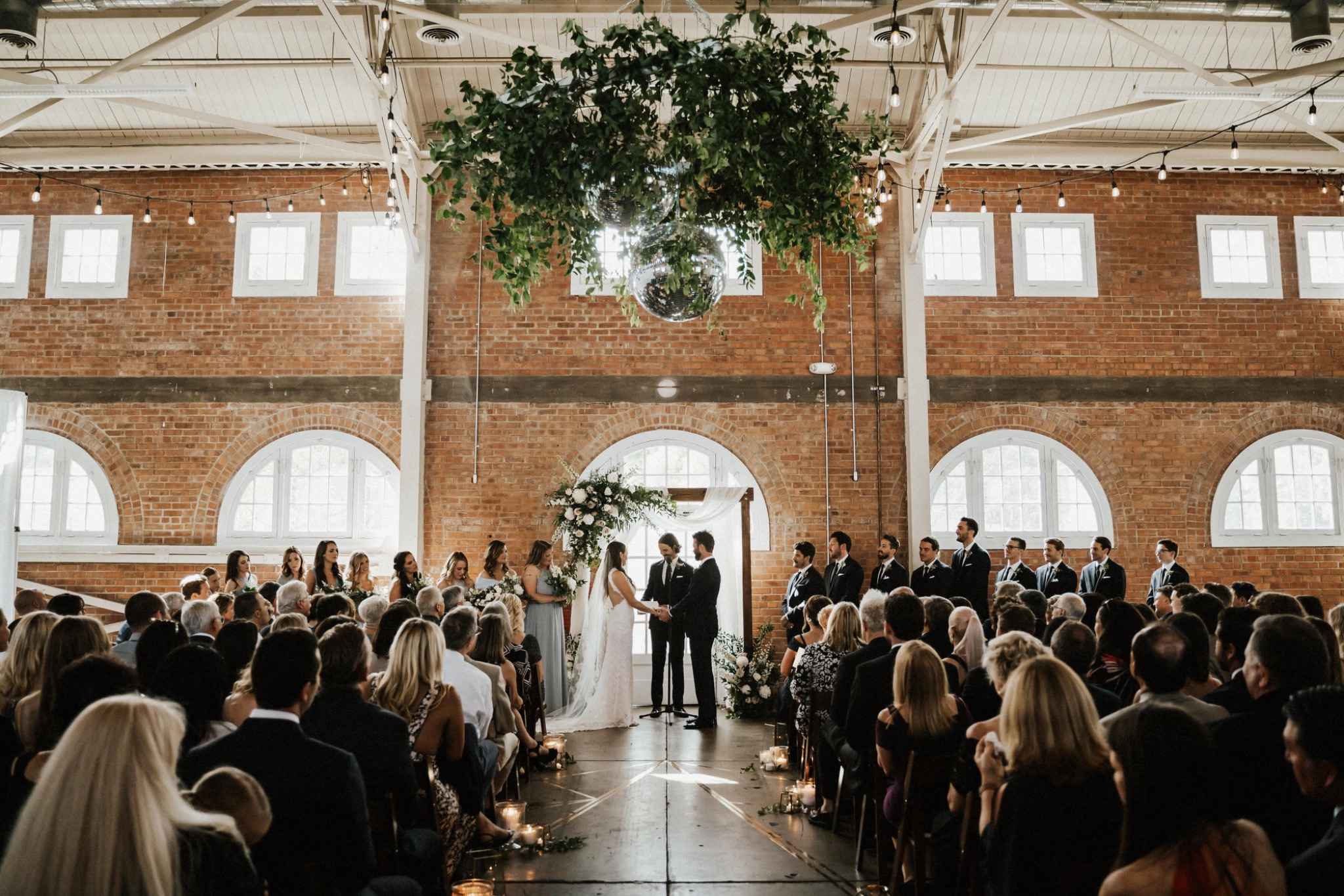 Indoor Minneapolis wedding ceremony featuring lush greenery arch and brick walls