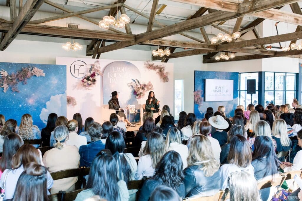 Women on stage speak to a crowd at a conference in large room with wooden beams and big windows