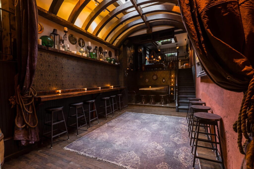 Moody, warm New York City bar with a curved ceiling, dark wood tables and chairs and velvet drapery