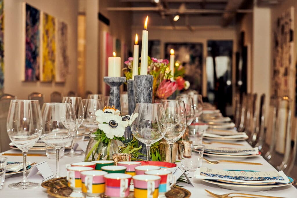 A closeup of a long banquet table decorated with flowers, glassware and colored cups in a gallery event space