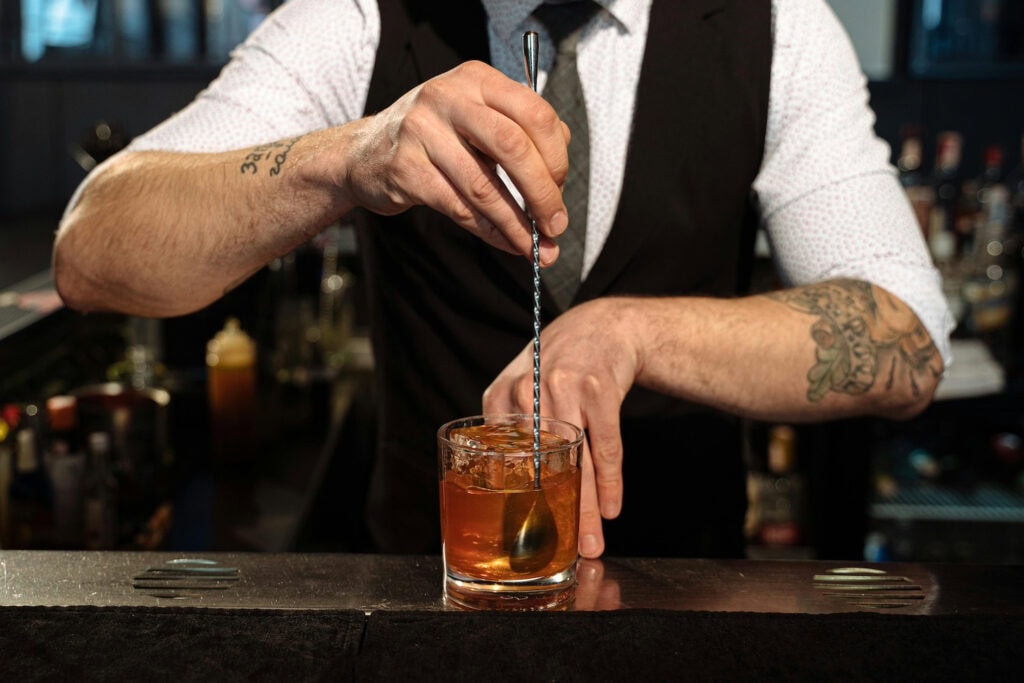 A bartender in a vest and tie stirs a Manhattan cocktail