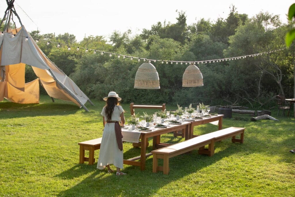 A woman standing at the end of a long wooden table outdoors in the sunshine beneath string lights and rattan hanging pendants next to a tent