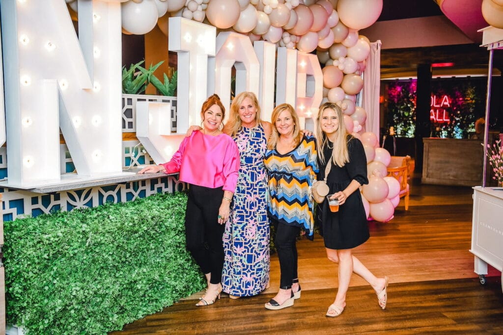Four women stand smiling by a large Bon Jour sign by a pink balloon arch