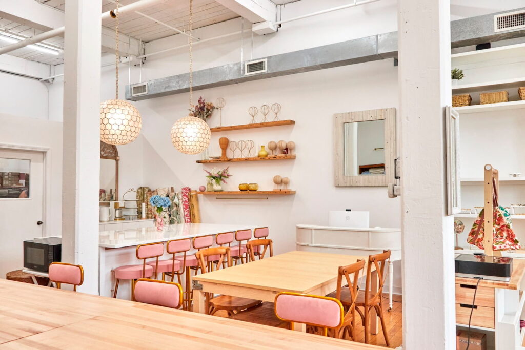 Pink and gold barstools and wooden chairs create different seating arrangements in a bright studio space with tall ceilings and white walls