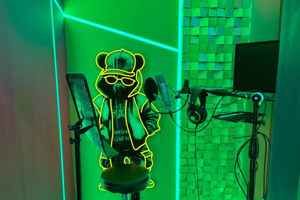 Green neon lights illuminate a small sound booth set up to capture vocals.