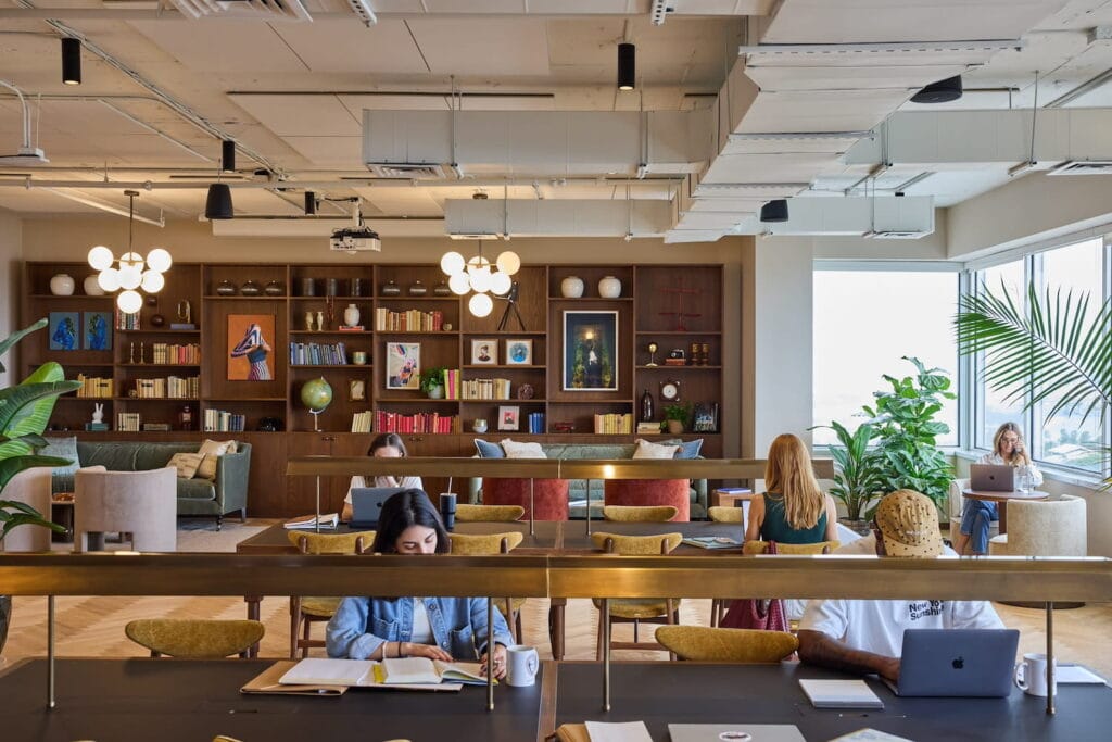 A warm coworking space with long tables, soft lighting and multiple seating areas in an office space with bright windows