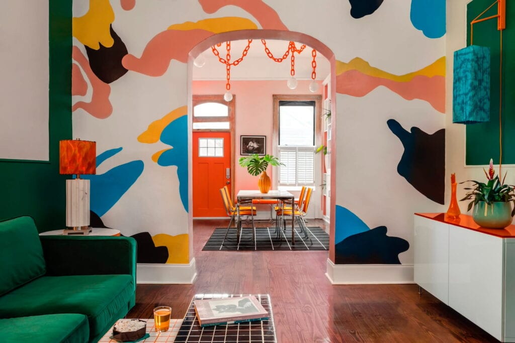 A brightly-colored interior of a New Orleans home with an orange front door, primary colored-walls and a green velvet couch