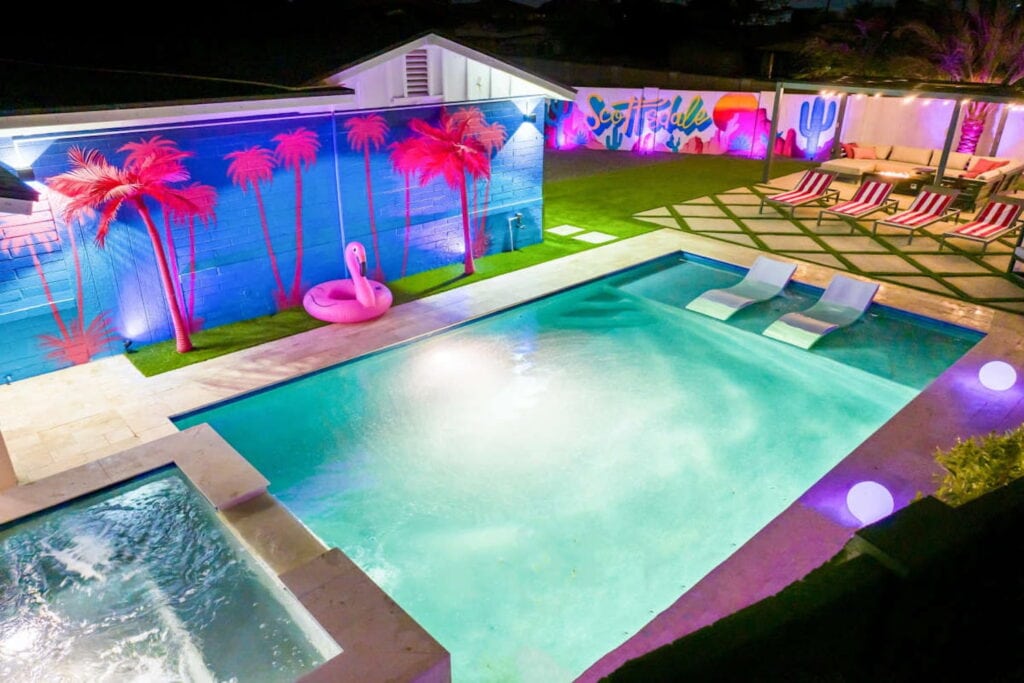 Ariel shot of a pool and hot tub illuminated at night and surrounded by a mural of pink palm trees, lounge chairs and a pink flamingo float