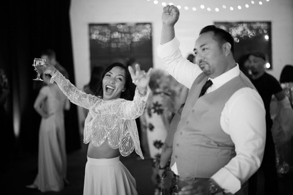 A bride holding a wine glass and dressed in a two-piece dress celebrates next to a man in a vest and tie