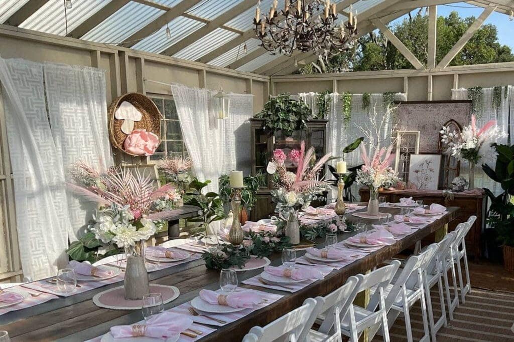 Greenhouse with chandeliers and a long wooden table set for a reception with white and pink linens and flowers