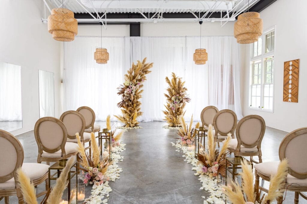 A bright white room illuminated by large windows draped in gauze is set up for an intimate wedding ceremony with bohemian style decor
