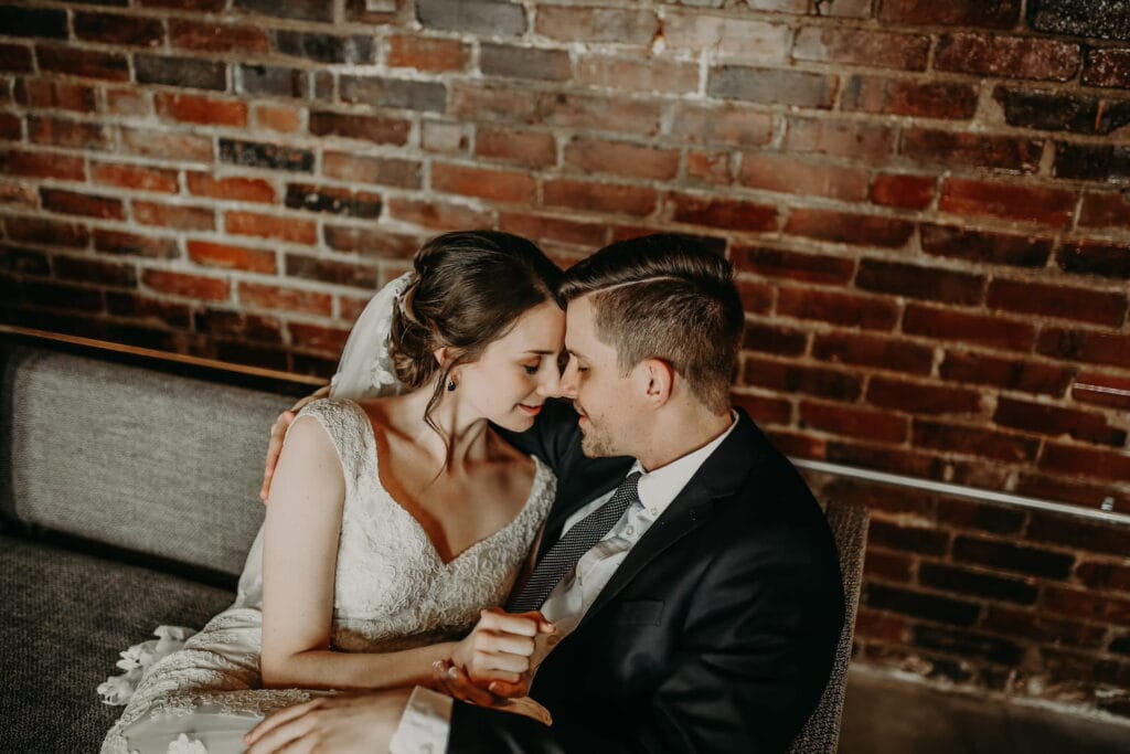 A bride and groom share a private moment on a couch by a brick wall